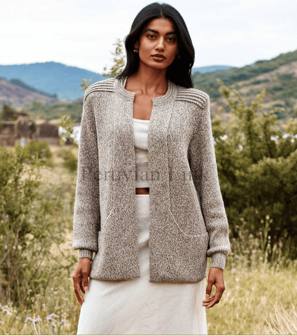 Woman wearing a textured cardigan over a white outfit in a natural setting with mountains and greenery.