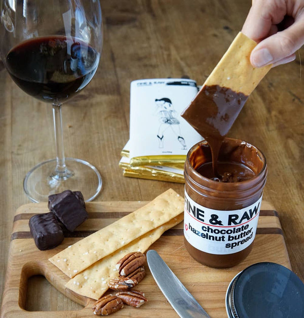 Person spreading chocolate hazelnut butter on a cracker with a glass of red wine and a jar of the spread on a wooden board.