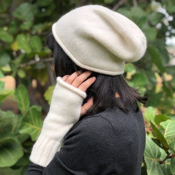 Person wearing a white knit beanie and gloves in front of green foliage