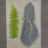 Gray textured napkin with string tied around it next to a green fern leaf on a striped fabric background