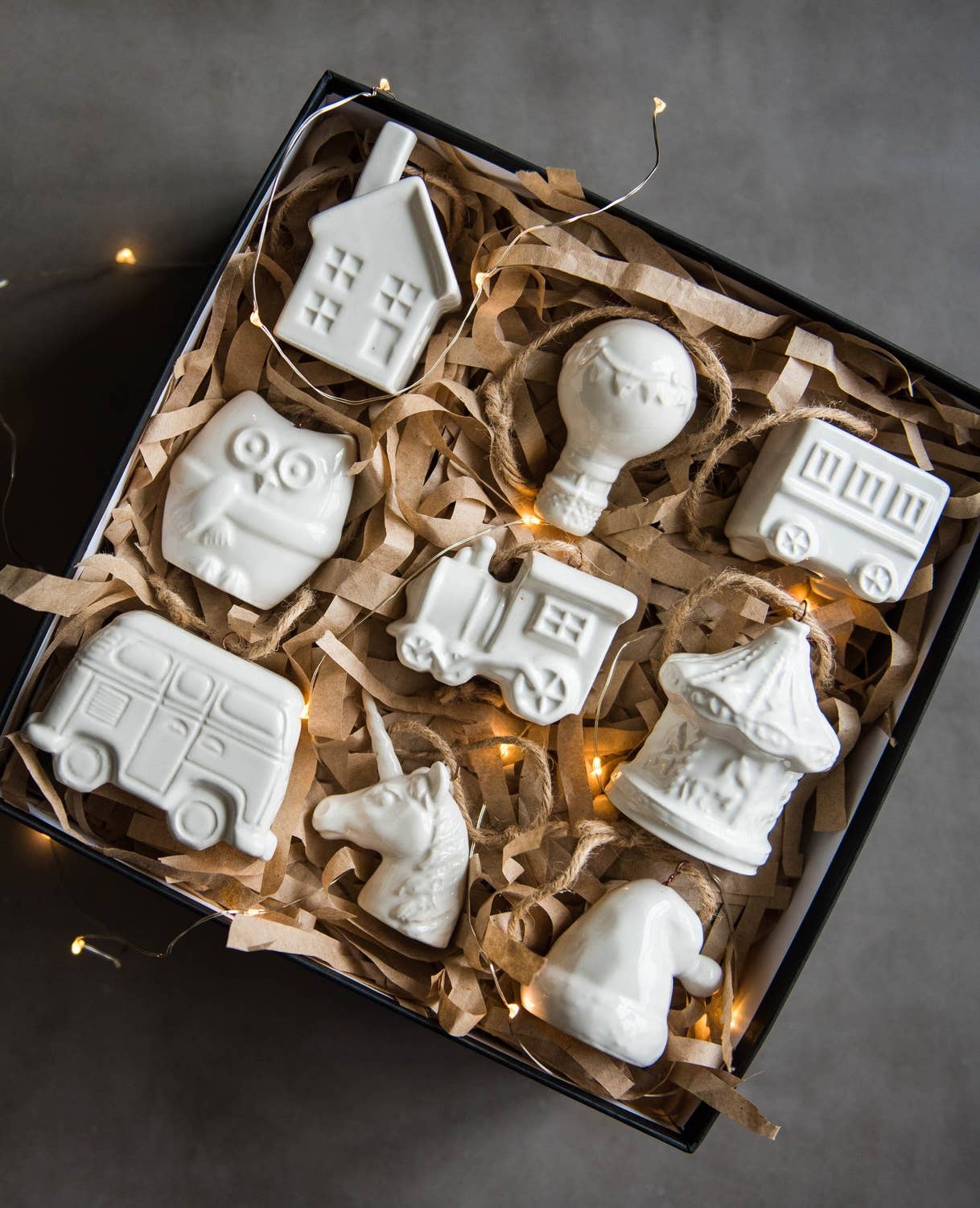 Set of white ceramic ornaments shaped like animals and vehicles in a box with brown tissue paper and string lights.