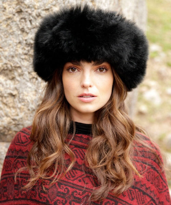 Woman wearing a black fur hat and red patterned sweater against a stone wall.