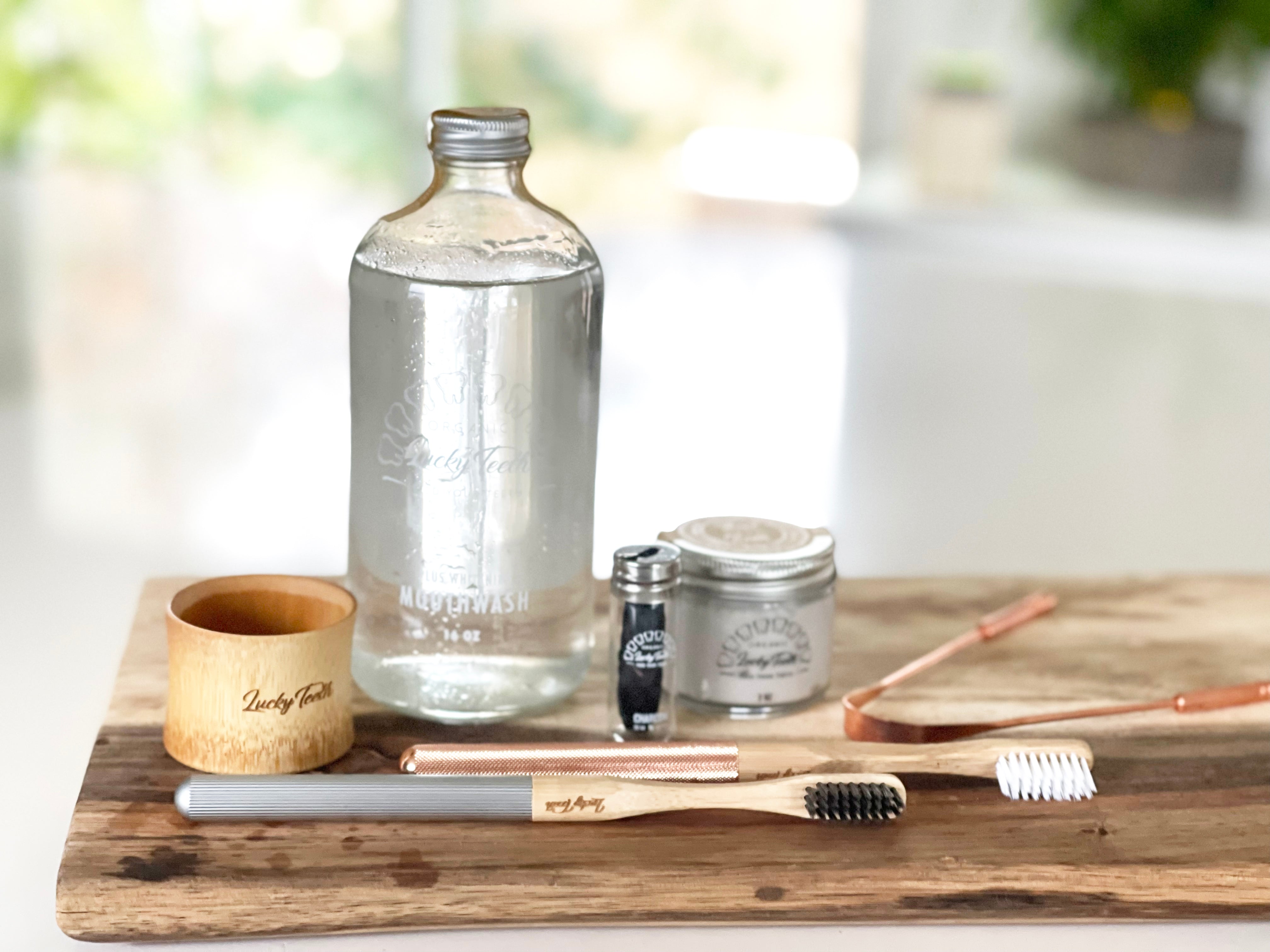 Zero-waste products including a glass bottle, bamboo toothbrushes, and wooden cup on a wooden tray.