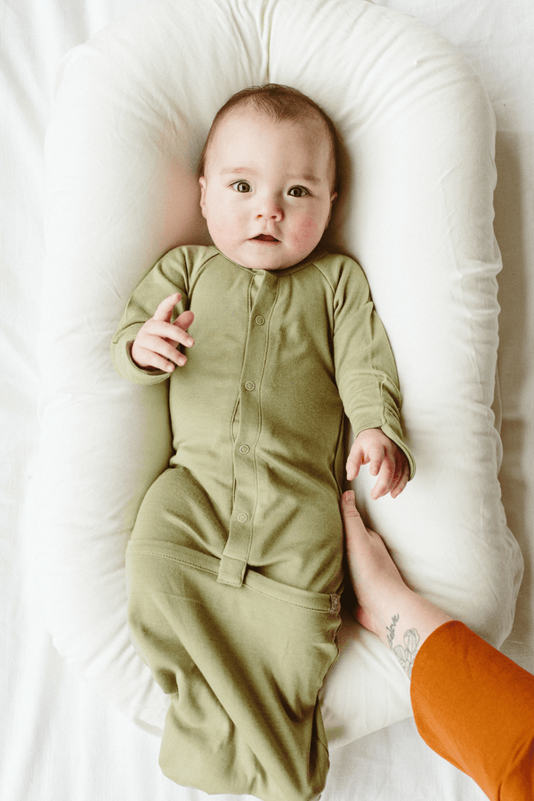 Baby in a green onesie lying on a white cushion with a person's foot visible.