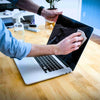 Person cleaning a laptop with a cloth on a wooden desk.
