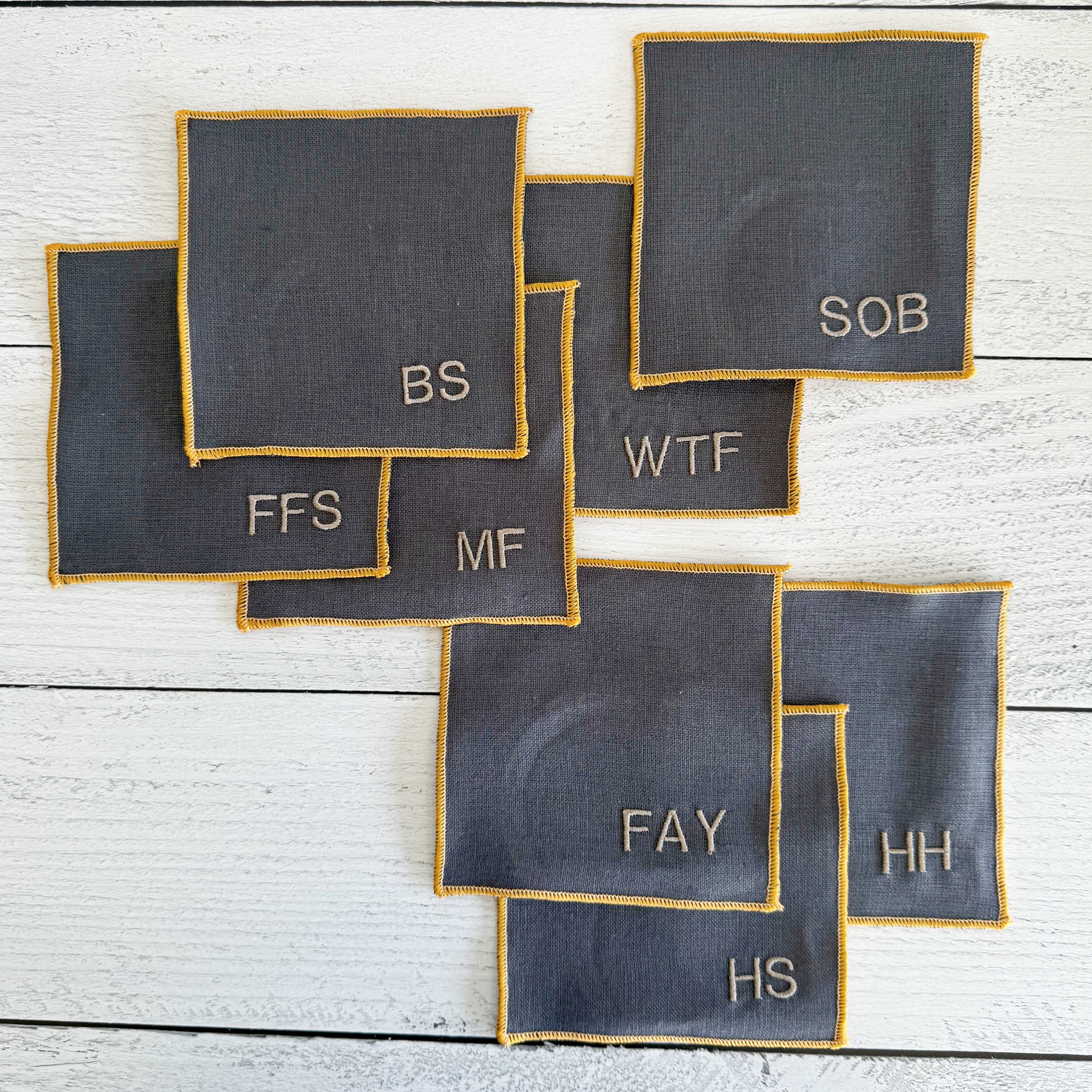 Close-up of grey linen sweary coaster showing yellow embroidered edging and letter design