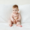 Baby in a pink outfit sitting on a white couch holding a wooden toy.