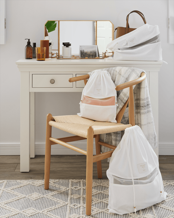 Three Hayden Hill organic cotton dust bags styled on a vanity table and chair in a bright bedroom