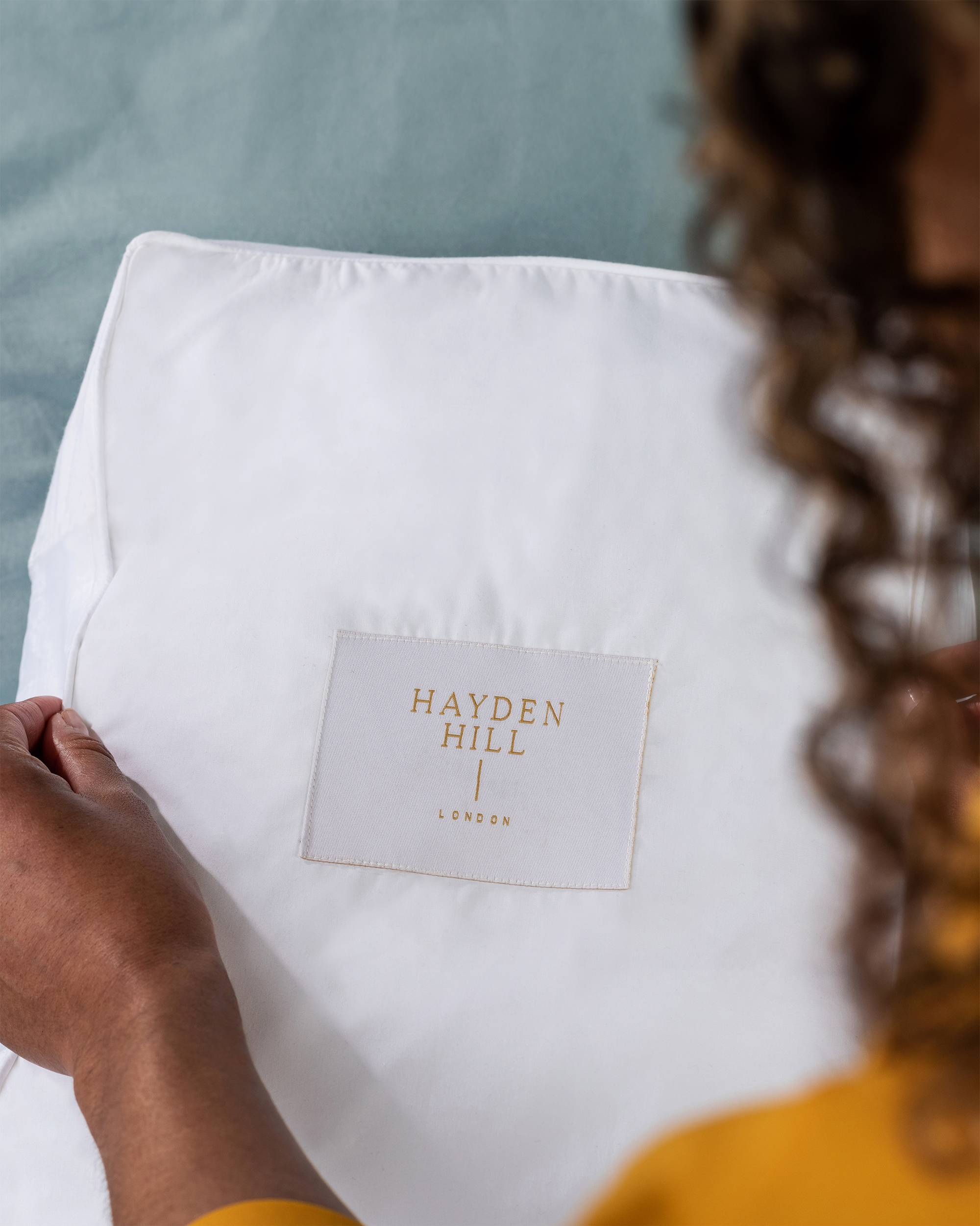Person holding a white pillow with 'Hayden Hill' label against a blurred background