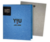 A blue eco-friendly greeting card with a red heart and the phrase 'YOU ARE HERE' printed on it, alongside a grey envelope.