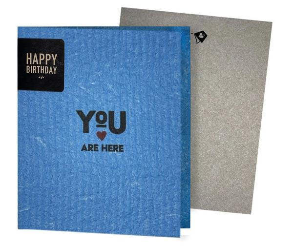 A blue eco-friendly greeting card with a red heart and the phrase 'YOU ARE HERE' printed on it, alongside a grey envelope.