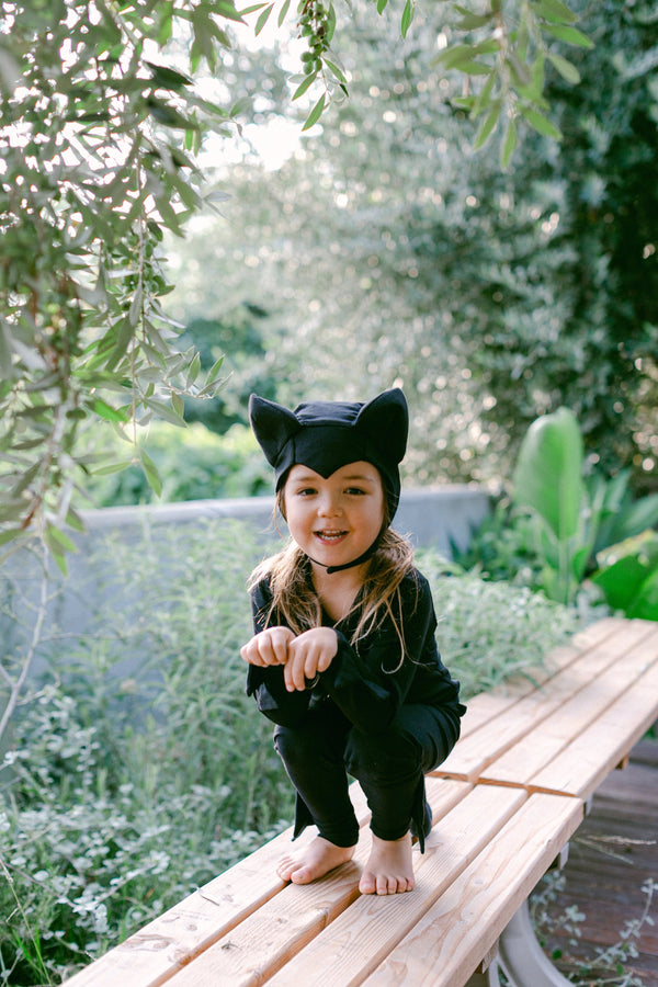Child in a black cat costume standing on a wooden bench outdoors.