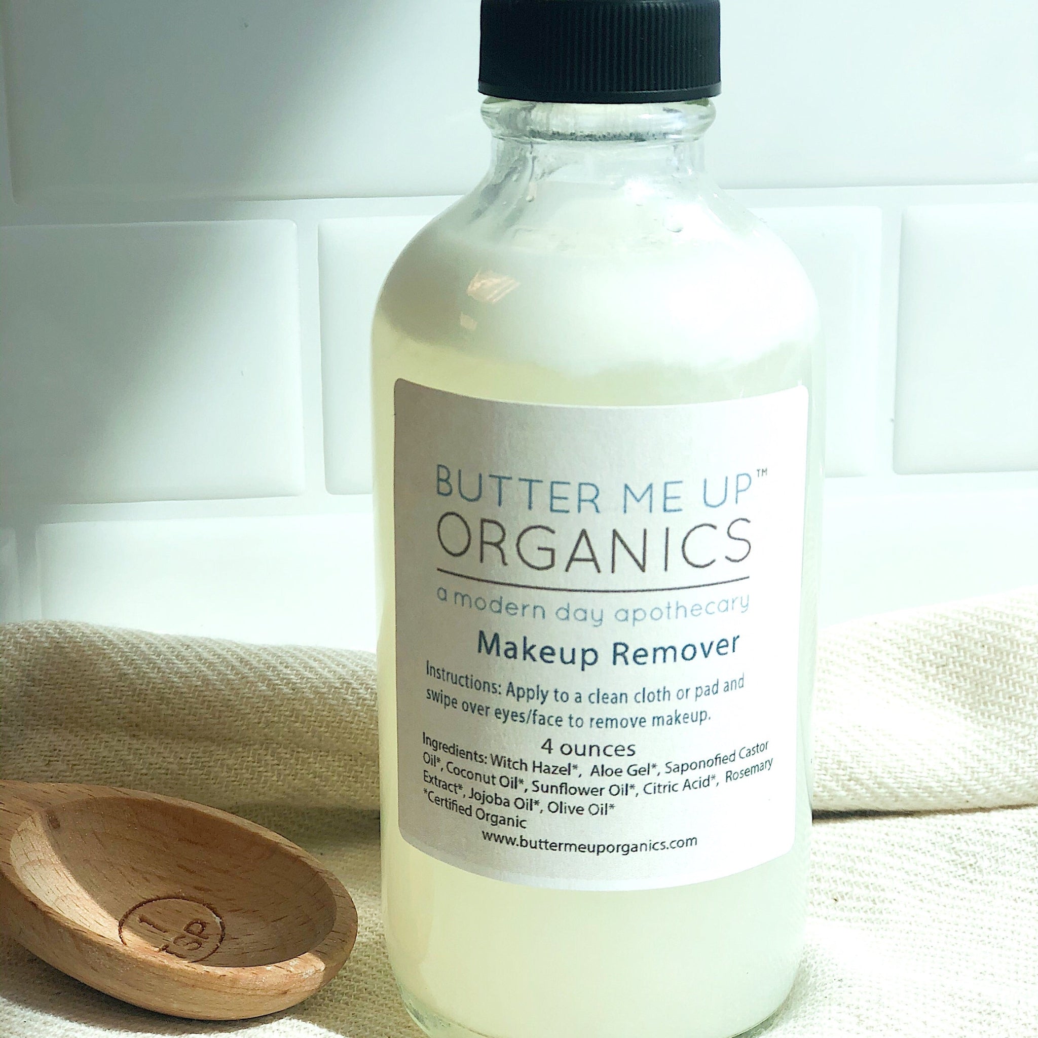 Butter Me Up Organics makeup remover bottle on a white surface with a wooden spoon and towel in the background.