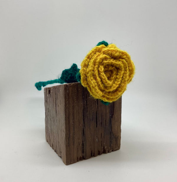 Yellow crocheted flower with green leaves on a wooden block against a white background