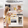 A children's play kitchen set in a bright room, with a young girl wearing a yellow striped apron and cap standing at the counter, and a young boy smiling while reaching into the kitchen. The kitchen features a white body with gold and yellow accents, including hanging utensils, a stovetop, oven, and storage cabinet.