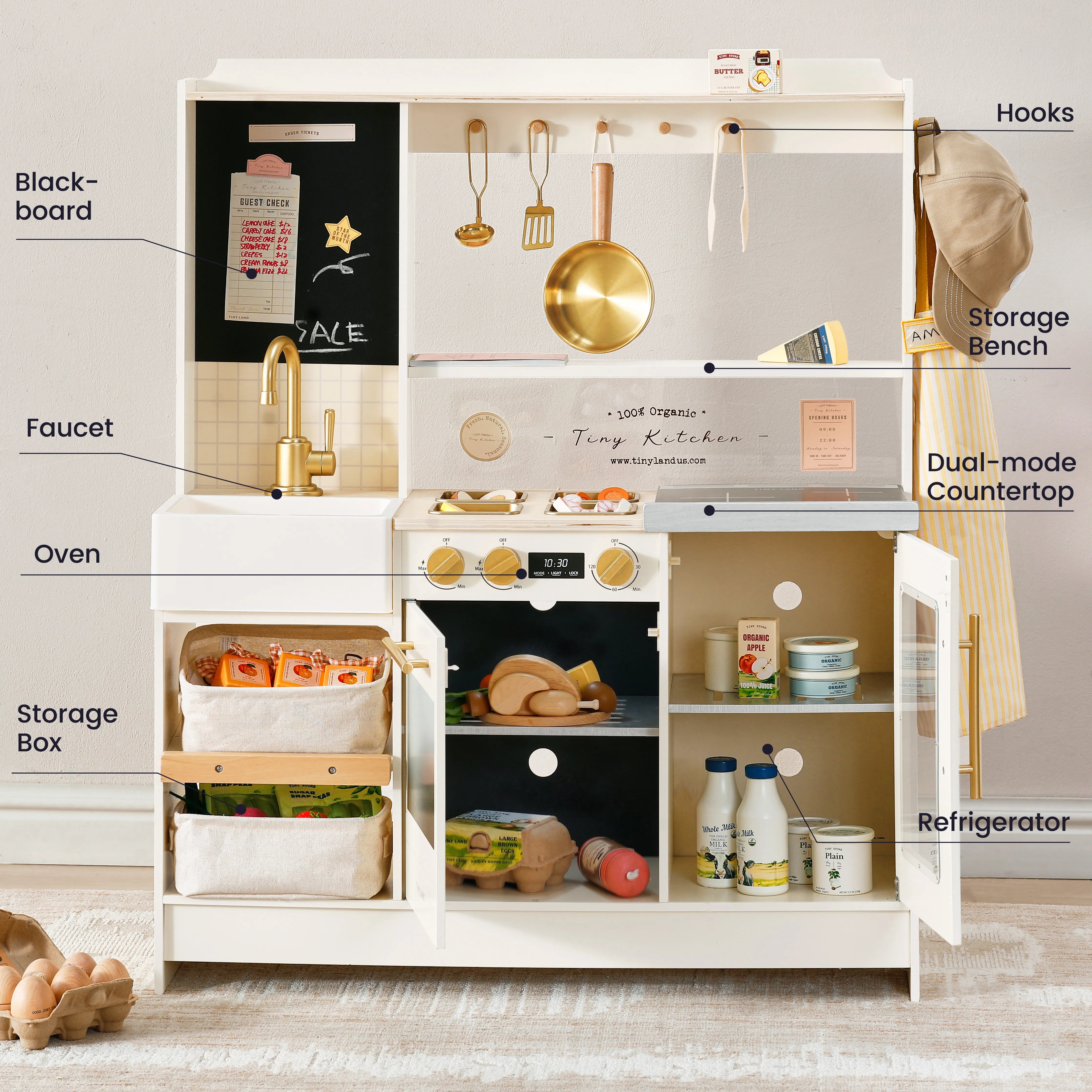 A children's play kitchen set with labeled callouts pointing to features such as black-board, faucet, oven, hooks, storage bench, dual-mode countertop, refrigerator, and storage box. The kitchen is cream-colored with gold-tone metal and brass details, styled with toy food items, utensils, and accessories inside an open refrigerator door.