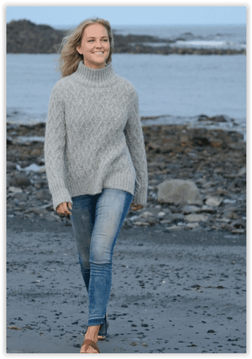 Woman wearing a gray sweater and blue jeans standing on a rocky beach.
