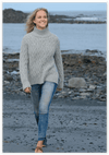 Woman wearing a gray sweater and blue jeans standing on a rocky beach.