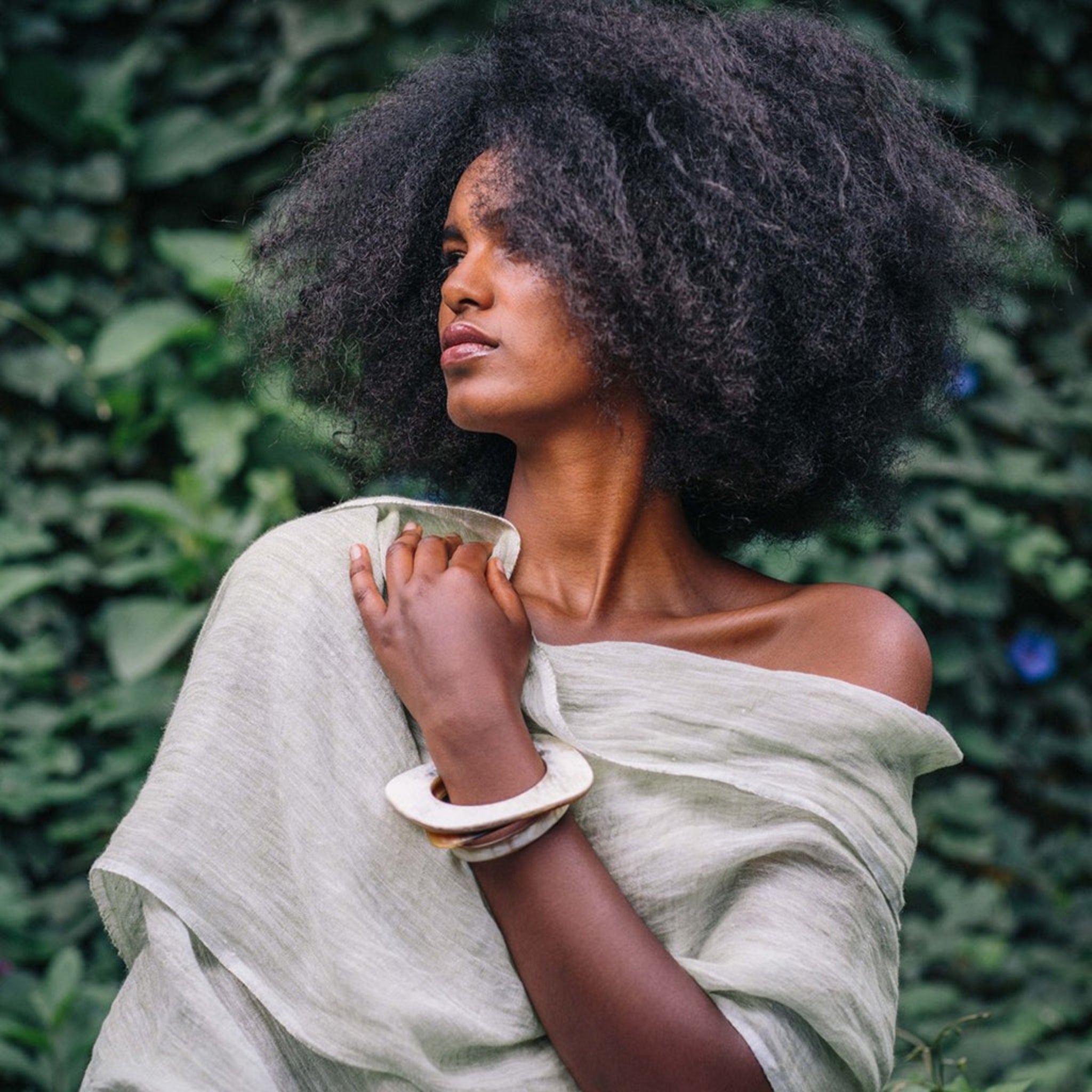 Woman with an afro wearing a light-colored off-shoulder garment against a green leafy background