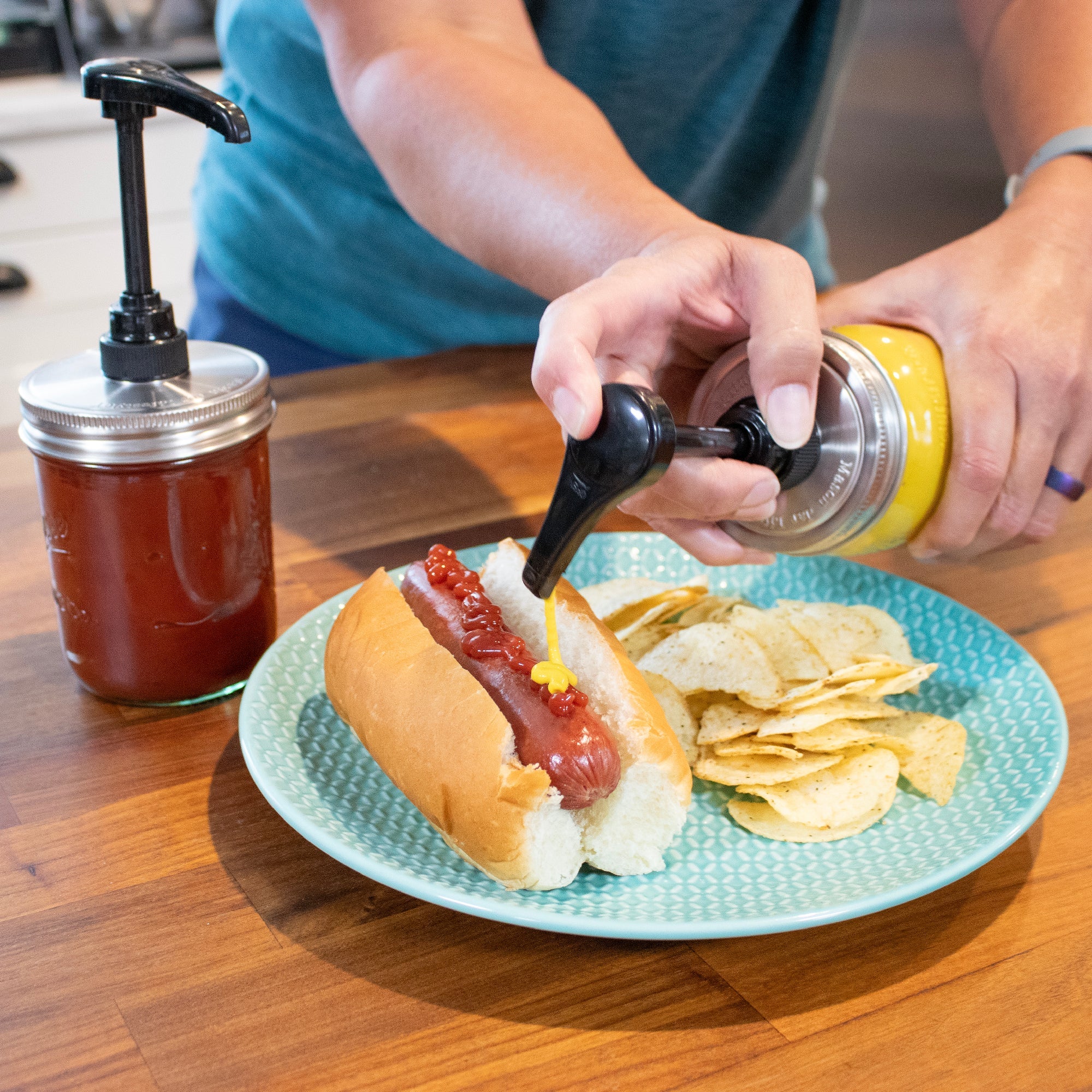 Person adding condiment to a hot dog on a plate with chips, next to a jar of ketchup.