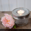 Glass jar with metal lid containing a lit candle next to a pink flower on a wooden surface. Transform your glass jars into glowing lanterns with the 6-Pack Tea Light Holder Metal Lid Inserts at Clozist. Rustic galvanized design leaves your jar free for decorative fill. The perfect sustainable gift for DIY decor, weddings, and cozy home lighting!