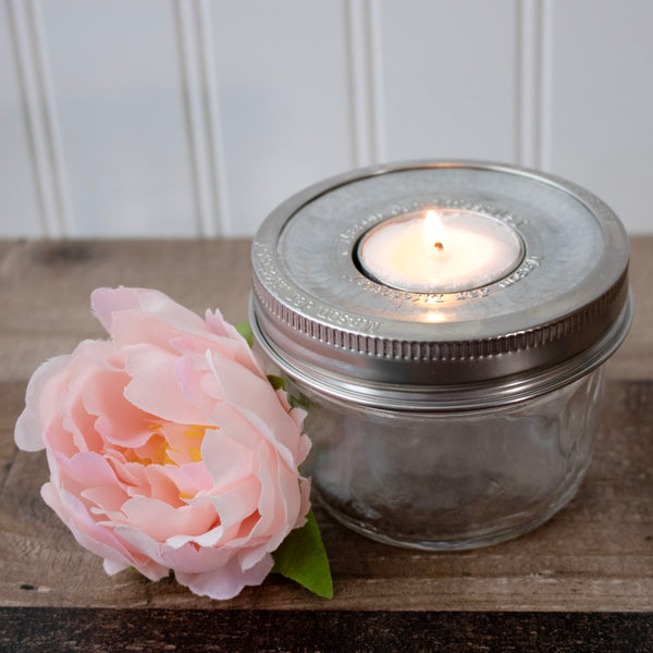Glass jar with metal lid containing a lit candle next to a pink flower on a wooden surface. Transform your glass jars into glowing lanterns with the 6-Pack Tea Light Holder Metal Lid Inserts at Clozist. Rustic galvanized design leaves your jar free for decorative fill. The perfect sustainable gift for DIY decor, weddings, and cozy home lighting!