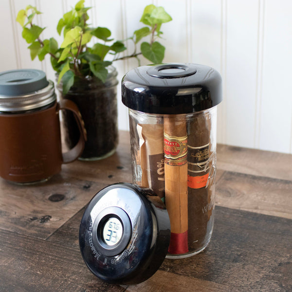 Jar with cigars and a digital thermometer on a wooden surface with a plant in the background.