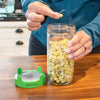 Person holding a jar of sprouts with a green sprout growth device on a wooden surface.