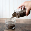 Hand pouring chia seeds from a jar into another jar on a wooden surface.