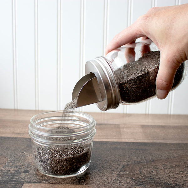 Hand pouring chia seeds from a jar into another jar on a wooden surface.
