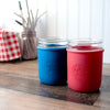 Two mason jar cups, one blue and one red, on a wooden surface with a checkered cloth and plants in the background.