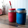 Red and blue mason jar cups with straws on a wooden surface.