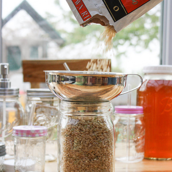 Person pouring grain into a glass jar with a metal funnel.
