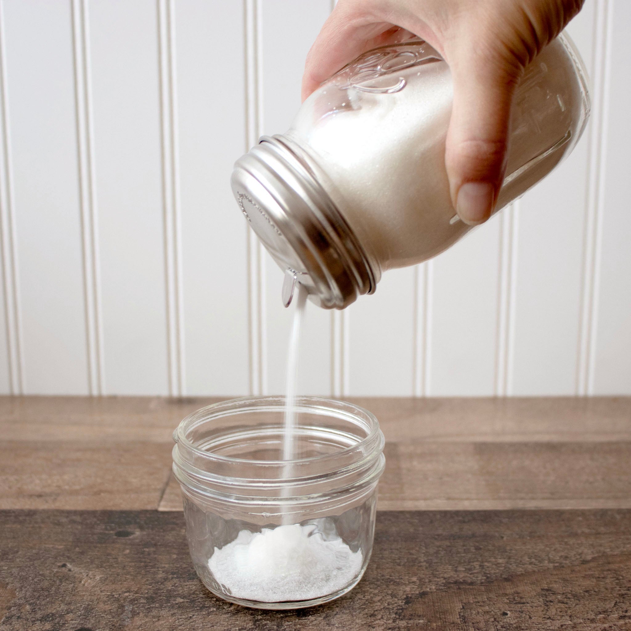 Hand pouring salt from a mason jar into another on a wooden surface with a white paneled wall background. Transform your Mason jars with the Sugar Dispensing Stainless Steel Lids at Clozist. Rust-resistant, food-safe, and dishwasher safe. A durable 2-pack designed for easy pouring and sustainable kitchen organization. The perfect green gift for bakers and coffee lovers!