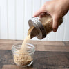 Hand pouring grain from a jar into another jar on a wooden surface.