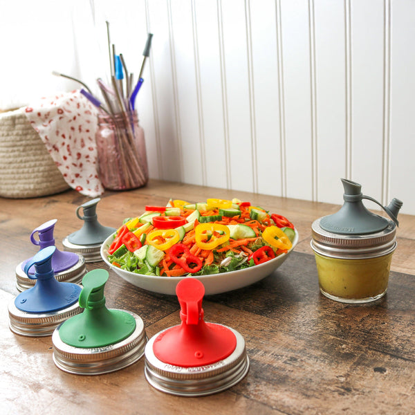 Colorful jar lids on a wooden surface with a bowl of salad in the background. Transform your Mason jars with the Silicone Oil Cruet Pour Lid at Clozist. Enjoy mess-free, non-toxic pouring for oils and dressings. Durable, dishwasher safe, and eco-friendly. The perfect sustainable gift for an organized, plastic-free kitchen!
