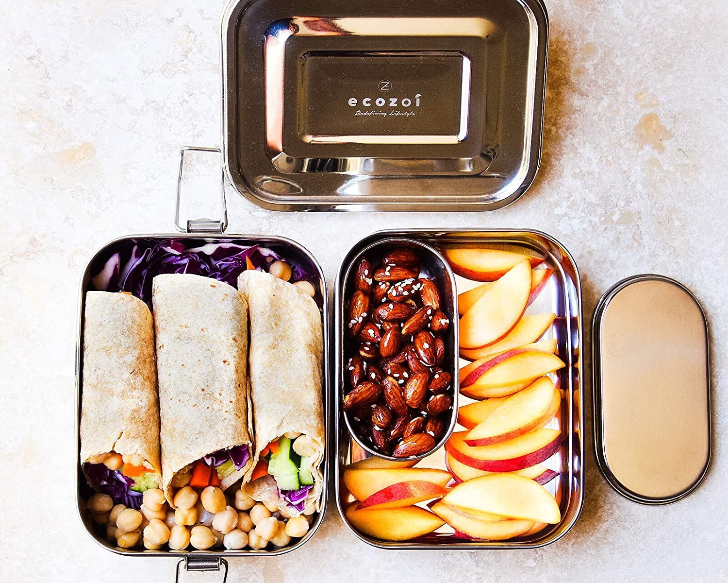 Metal lunch box with compartments containing wraps, fruits, and nuts, branded 'ecozoi'.