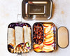 Metal lunch box with compartments containing wraps, fruits, and nuts, branded 'ecozoi'.