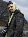 Man wearing a black puffer jacket with a brand logo, holding a camera, standing against a mountainous background.