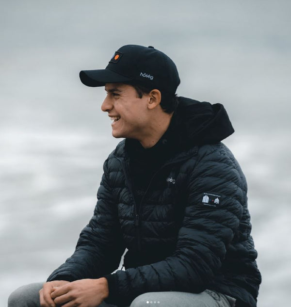 Person wearing a black cap and jacket sitting outdoors with a cloudy sky.