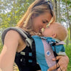 Woman holding a baby in a carrier outdoors with trees in the background