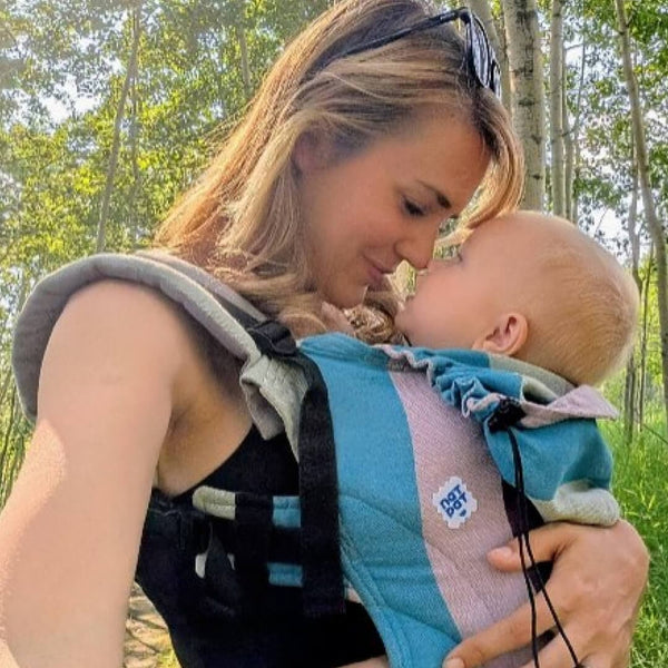 Woman holding a baby in a carrier outdoors with trees in the background