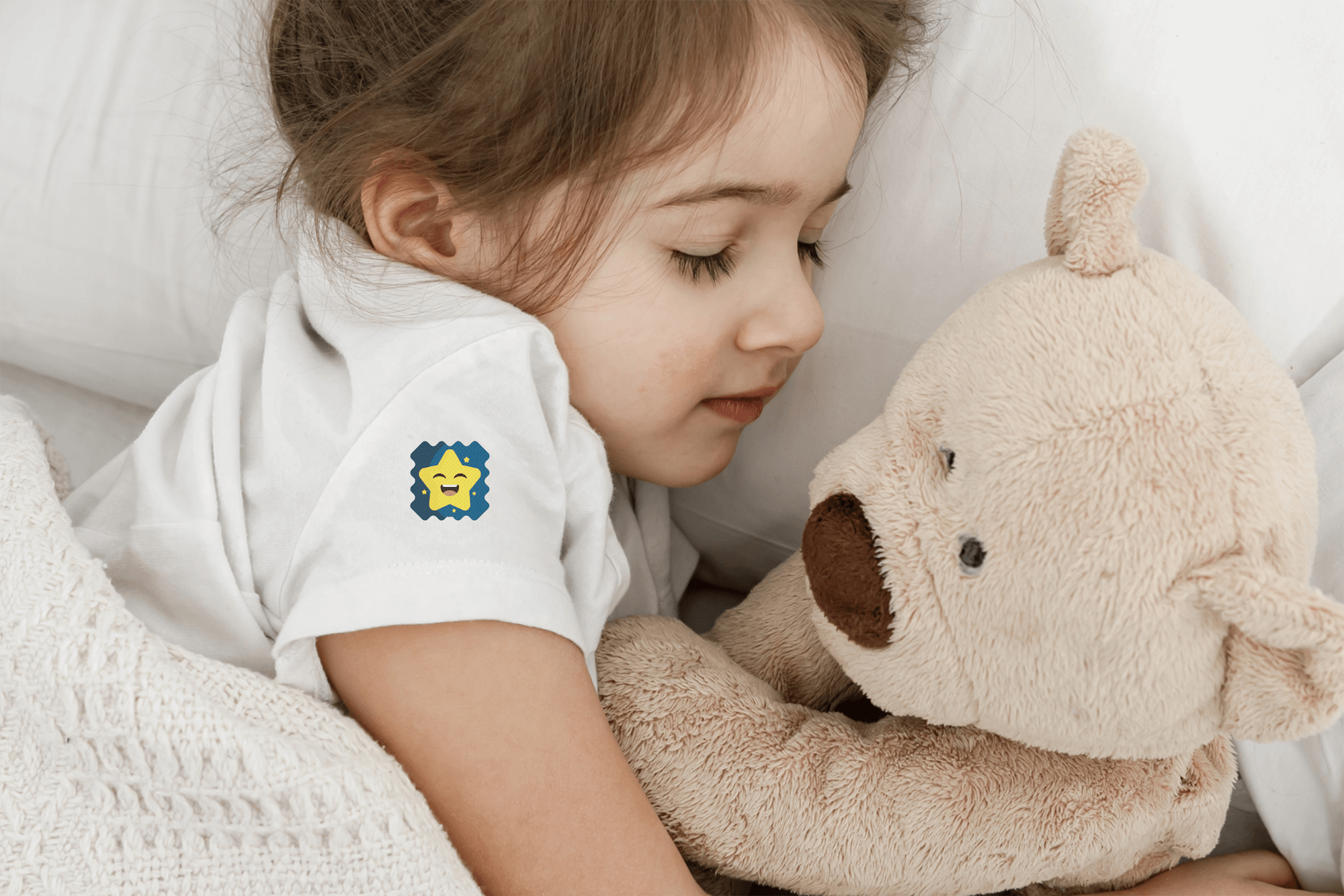 Child holding a teddy bear wearing a white shirt with a logo, sitting on a white blanket.