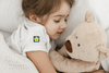 Child holding a teddy bear wearing a white shirt with a logo, sitting on a white blanket.