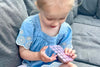 Child playing with a small pink keyboard on a gray couch. SunnyPatch UV stickers change color when it's time to reapply sunscreen — safe for babies & kids. Fun, smart sun protection for beach, pool & outdoor play. Shop now.
