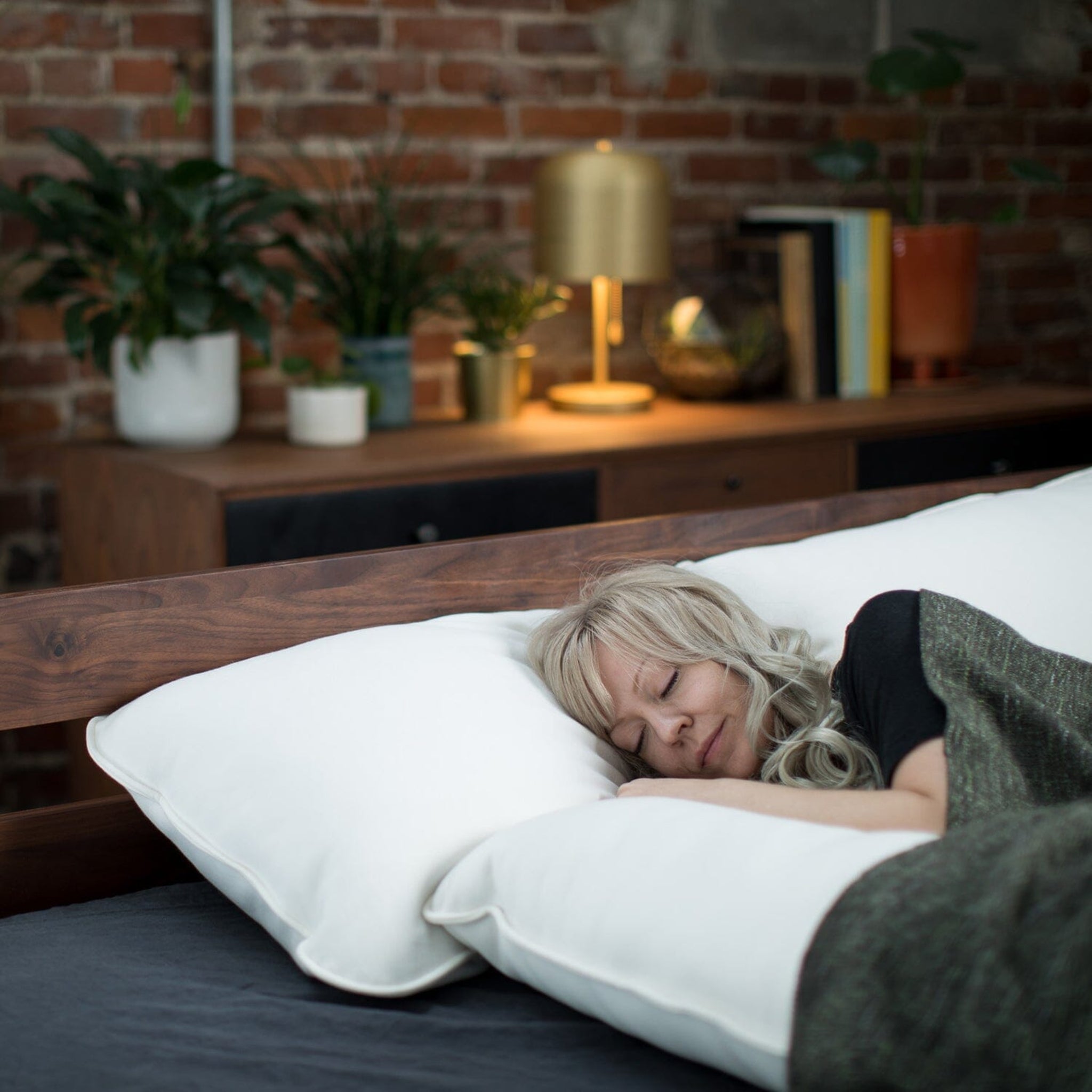 Woman sleeping on a bed with white pillows in a cozy room with plants and a lamp.