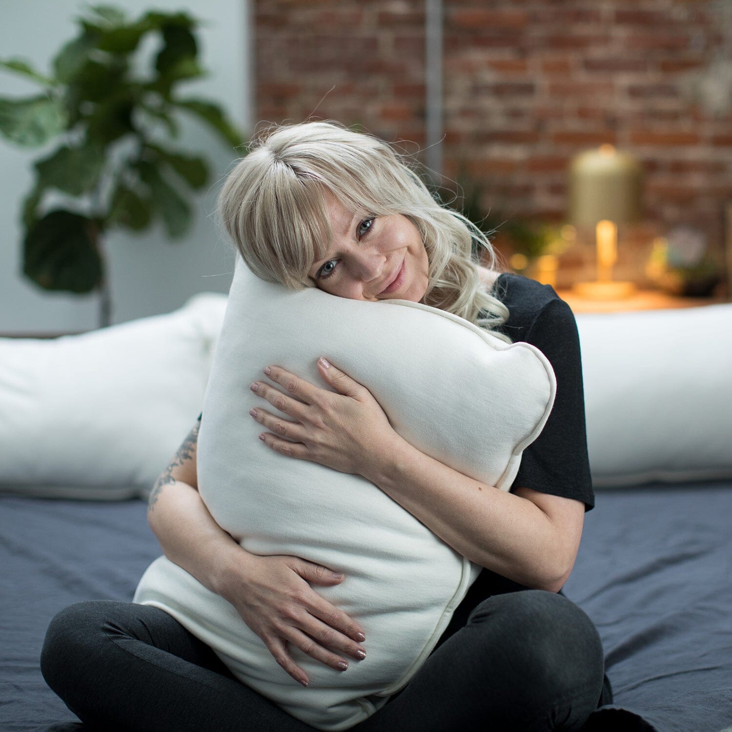 Woman hugging a large white pillow in a cozy room with a brick wall and plants.