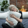 Woman hugging a large white pillow in a cozy room with a brick wall and plants.