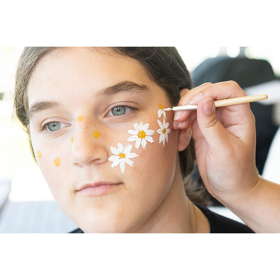 Person applying white flowers with yellow centers to a person's face using a brush.