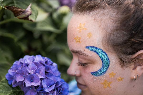 Person with face paint featuring a blue crescent moon and yellow stars, holding purple flowers.